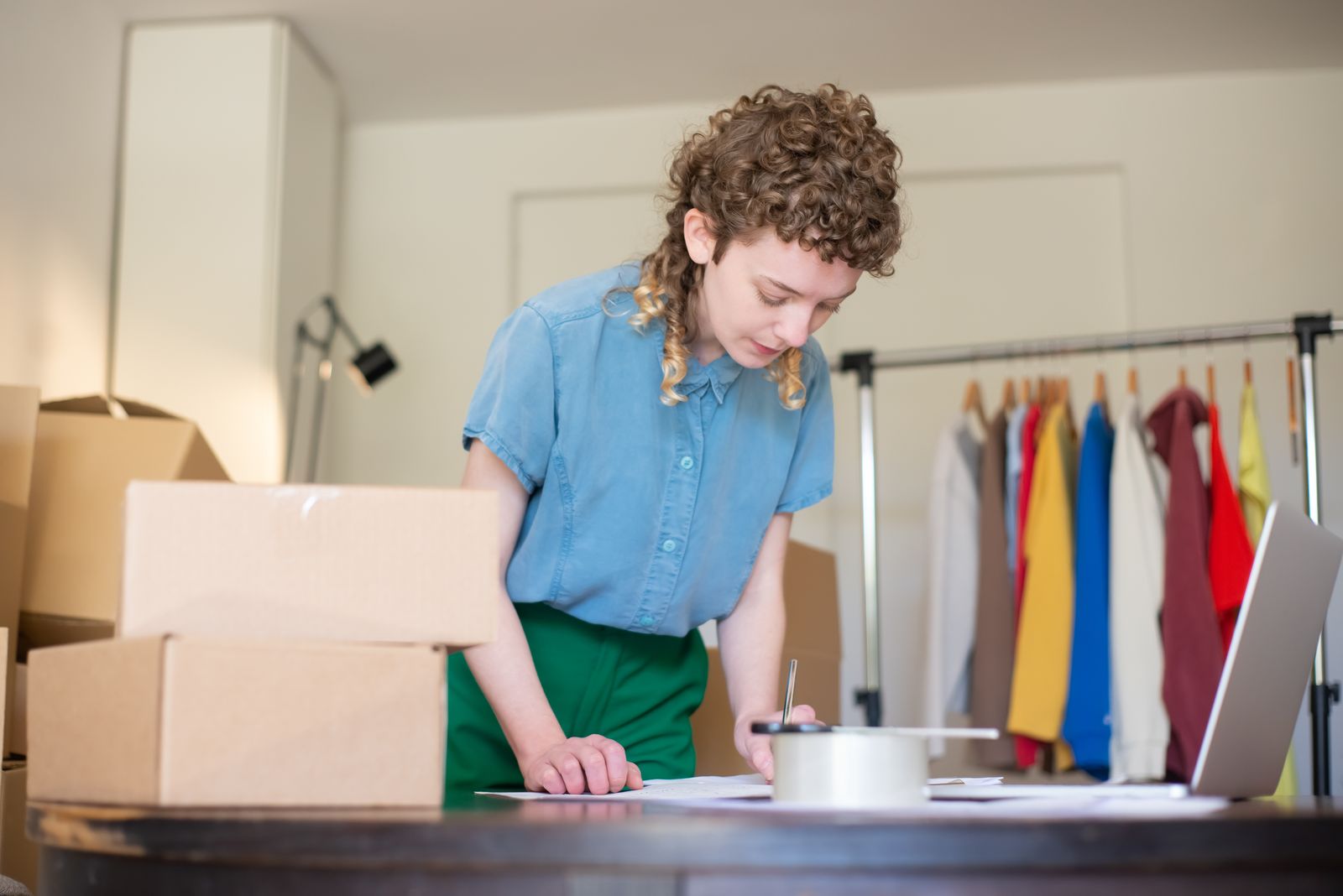 Mujer preparando pedidos de su tienda online junto a un portátil, cajas y un burro de ropa